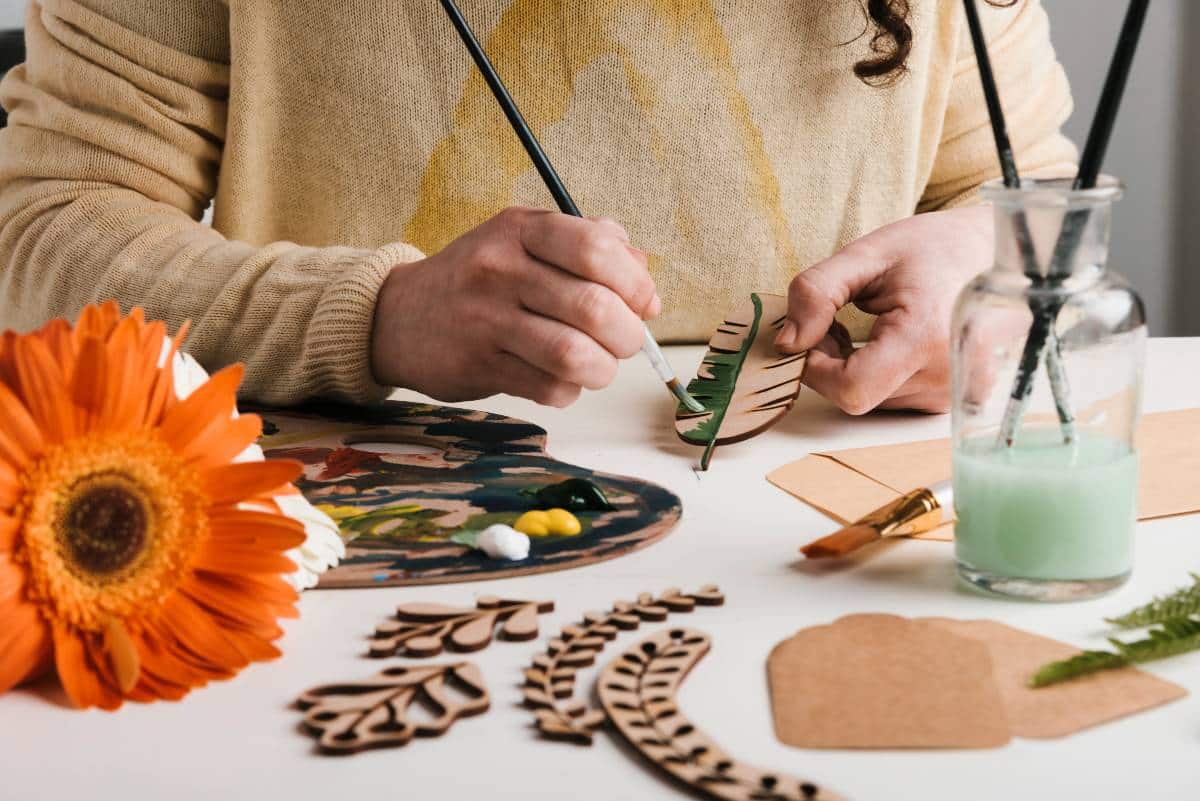 Mãos pintando objeto sobre mesa com diversos materias de artesanato. 