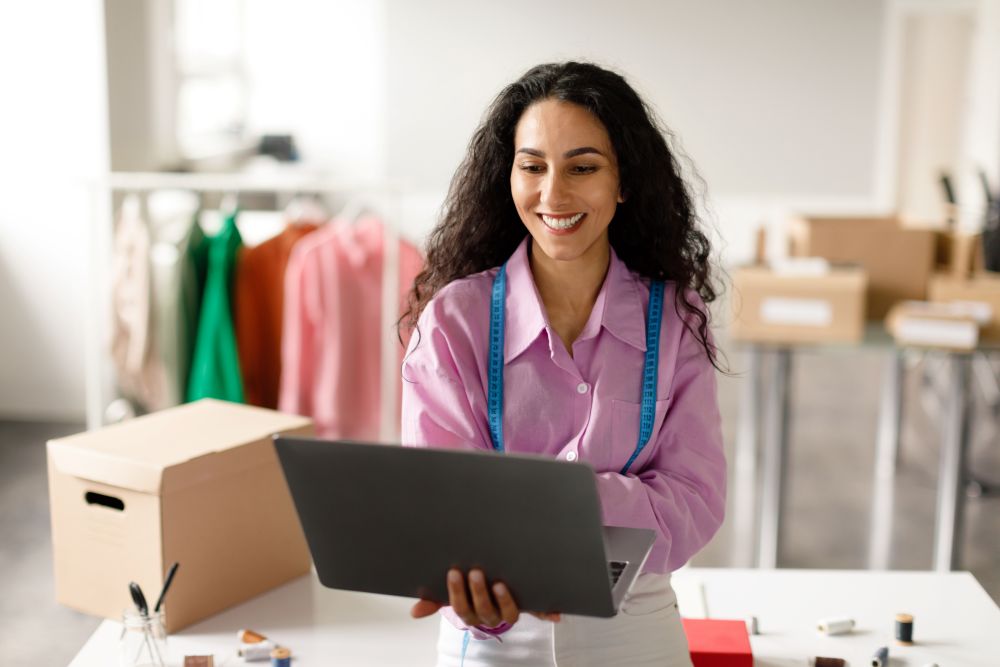 Mulher empreendedora usando notebook em loja de roupas com caixas de envio, gerenciando pedidos de e-commerce