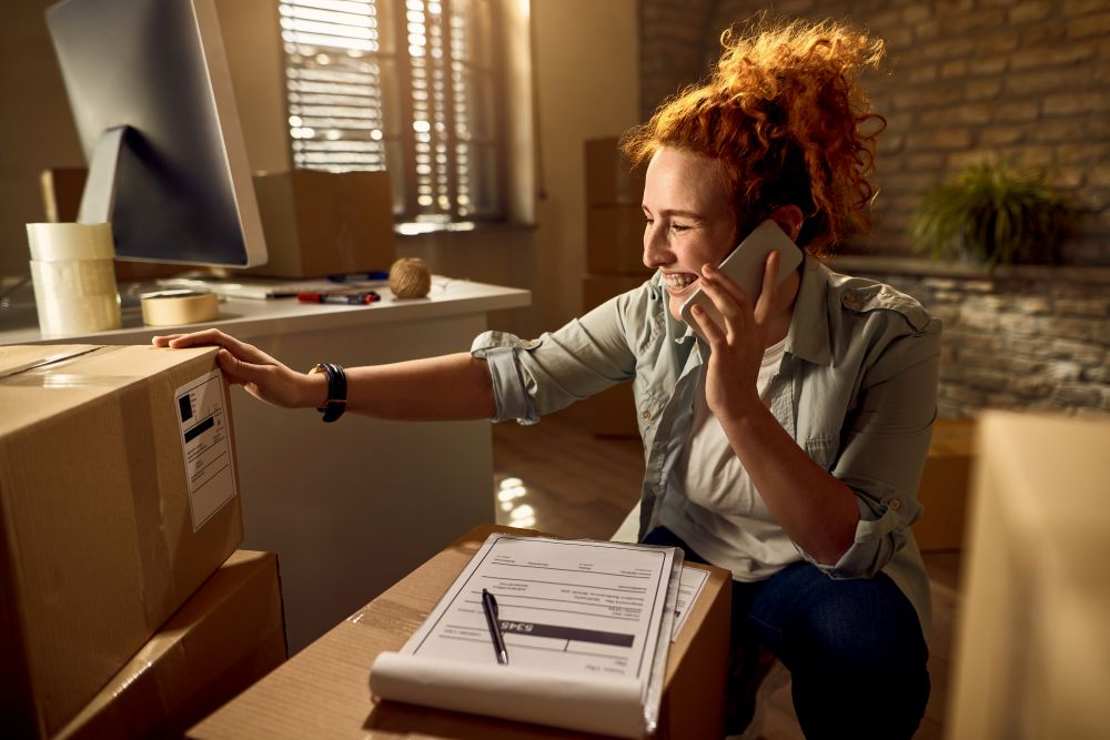 Empreendedora organizando pedidos de dropshipping em loja virtual enquanto conversa ao telefone e prepara envio de produtos de e-commerce.