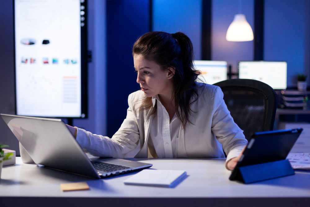 Mulher estudando cursos de marketing digital à noite, usando notebook e tablet em ambiente de trabalho moderno.