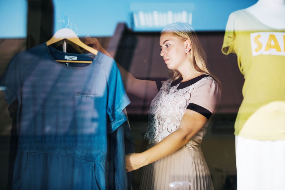 Mulher organizando vitrine de loja de roupas com peças de fast fashion, vista através do vidro.