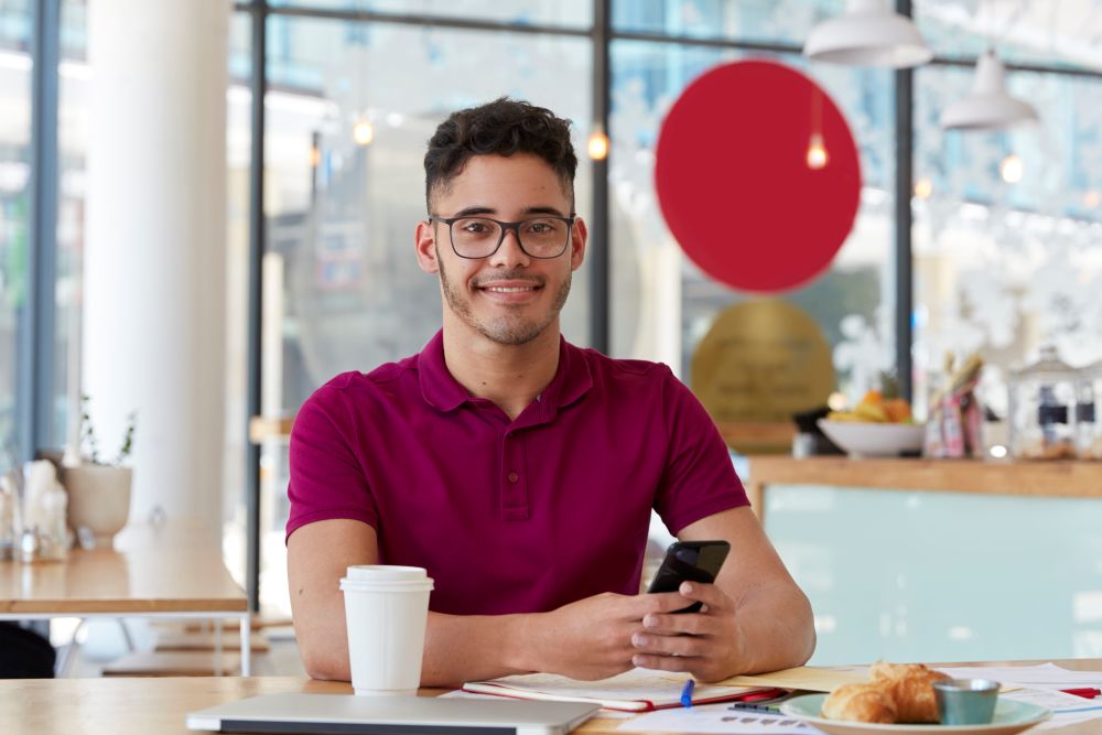 Jovem empreendedor sentado em cafeteria com celular na mão, representando MEI buscando linha de crédito para impulsionar seu negócio.