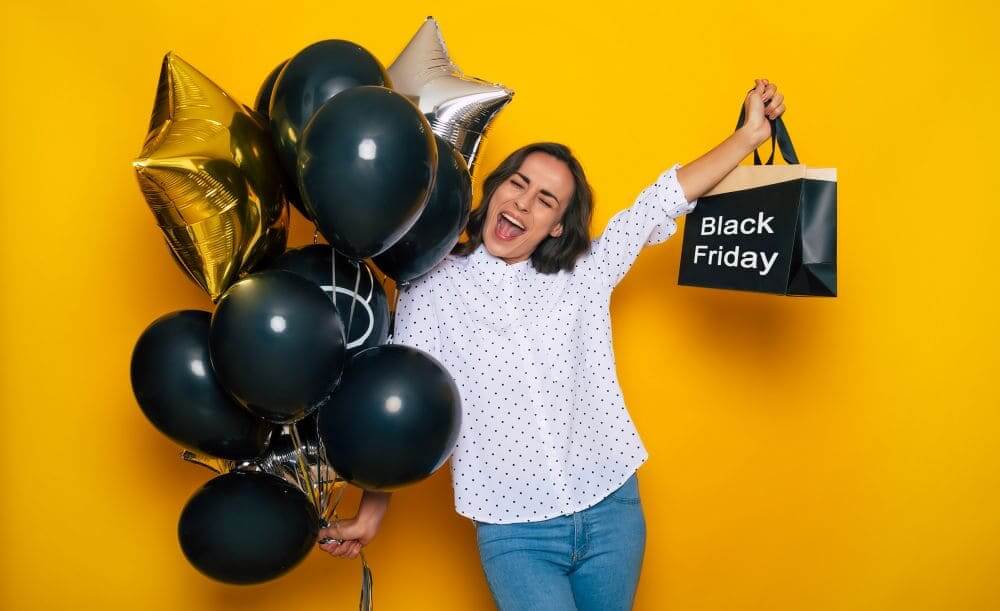Mulher animada segurando sacola escrita Black Friday e balões pretos e dourados, simbolizando compras e promoções da Black Friday