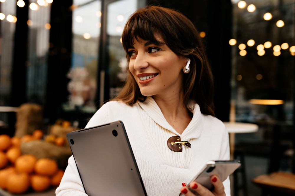 Mulher sorrindo ao ar livre segurando notebook e smartphone, representando mobilidade e praticidade do modelo SaaS para negócios digitais